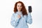 Skeptical redhead girl look confused, pointing at empty mobile phone screen with doubtful face, standing upset with app