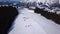 Ski resort in sunny weather. Snow on the ski slope of a wooded mountain. Many tourists near the ski lift station and cafe. Aerial