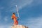 A skier-athlete standing on the edge of a high rock balances his skis in his hands against the blue sky and cirrus