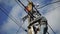 Skilled electrician fixes wires standing on ladder near high pole against blue sky on summer day backside view