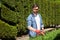 Skilled male gardener in protective glasses cutting greenery with hedge trimmer in topiary park.