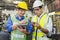 A skilled mechanic in a metal lathe is checking parts on a tablet. Engineers are working and repairing machines in industrial