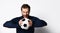 Skillful soccer player in blue tracksuit is smiling and holding a ball, posing isolated on white studio background. Close up