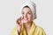 Skin care and beauty treatments concept. Positive young female uses cotton disk for applying cosmetics on face, wears towel and