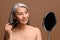 Skin care routine. Adorable middle-aged 50s Asian woman with long gray hair and naked shoulders holding mirror and