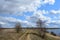 Sky, clouds. Winding road. River. Trees, shrubs, grasses on the low banks of the river. Forest in the distance. Rural view