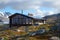 A sleek, contemporary cabin with large windows sits in a remote mountain landscape, offering a view of a stunning glacier and