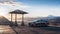 A sleek silver car is parked next to a wooden shelter, offering stunning views of mountains in the distance