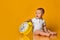 Sleepy infant baby boy toddler is sitting at holding big yellow alarm clock on yellow background with copy space