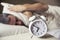 Sleepy young man covering ears with pillow as he looks at alarm clock in bed.