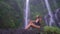 Slender adult girl in black bathing suit sits under great tropical cascade, admiring tropical nature