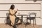 Slender black haired female sits at table outdoors, having cup of hot drink, relaxing during free time, taking photos of urban