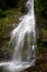 A slender blonde stands under a large picturesque waterfall, spreading her arms to the sides