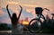 Slender girl sits with her back arms raised near retro bicycle with basket enjoying colorful sky with clouds at sunset