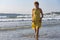 A slender lady in a yellow sundress walks on the water against the background of the oncoming sea wave.
