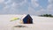 A slender man on the beach is flying a multi-colored kite. Active recreation on the banks of the river.