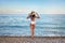 A slender tanned brunette in a swimsuit and a straw hat stands on a pebble on the ocean with her back to the camera