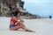 Slender tanned woman in colored swimsuit sits on ocean beach with white sand