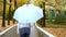 Slender woman in white leather jacket and jeans walking in the autumn park with a blue umbrella in the rain