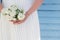 Slender woman in white satin pleated dress holding a small bouquet of white flowers in hands on a blue lath background with