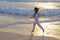 Slender woman in a white tracksuit jogging along the edge of the waves on the sandy beach of the sea