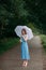 A slender young girl with blond long hair, in a blue summer dress and shoes, stands with a white umbrella