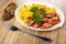 Slice of bread, white dish with slices of fried potatoes, parsley, sausage, fork on wooden table