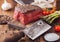 Slice of Raw Beef Topside Joint with Salt and Pepper on wooden chopping board with meat hatchet, garlic and asparagus tips