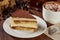 Slice of tiramisu cake on a plate with a cappuccino on a wooden table