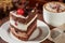 Slice of triple chocolate sponge cake with a cherry on top with a cappuccino on a wooden table