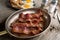 Slices of bacon frying in a skillet  with eggs and toast plated in the background