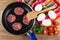 Slices of sausage in frying pan, eggs, spatula on napkin, bowl with parsley, halves of tomato on table. Top view