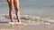 Slim female legs and feet standing on the sandy beach along sea water waves. Pretty woman walks at seaside surf. Girl