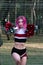 Slim fit girl cheerleader with pink hair in sportswear posing outdoors near fence at stadium with red and silver pompoms