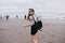 Slim pale girl with black backpack walking around beach with happy smile. Outdoor shot of magnificent lady having fun