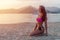 Slim tanned model in bikini posing on beach sitting sand in the light of morning at sunrise with mountains and sea in