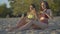 Slim tanned women drinking healthful juice as sitting on beach mat at sunset. Side view portrait of beautiful young