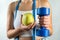 Slim unrecognizable woman with tape measure holding blue dumbbells and apple.