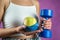 Slim unrecognizable woman with tape measure holding blue dumbbells and apple.