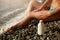 Slim woman tanned lower body in shape lying on pebble beach near sea waves and surf with sunblock cream bottle. Girl