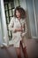 Slim young fashion model wearing white coat in window frame. Lovely fashionable woman with light brown curly hair