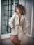 Slim young fashion model wearing white coat in window frame. Lovely fashionable woman with light brown curly hair