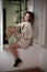 Slim young fashion model wearing white coat in window frame. Lovely fashionable woman with light brown curly hair