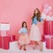 Slim young woman with trendy hairstyle posing with daughter in stylish attire next to pink presents with balloons and