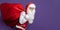 slinking santa claus carrying huge bag of gifts on purple background while looking at camera.