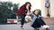 Slow motion of cheerful young women friends riding skateboard sitting on it and pushing it in the city on summer day