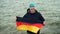 Slow motion portrait of joyful German sports fan holding flag of Germany and smiling while standing near the lake and