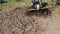 Slow motion rotary tiller blades loosen soil, ground scatters from cutter wheel.