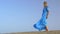 Slow motion view of young blond woman standing against blue sky in long blue dress and it is fluttering on the wind