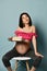 Sly pregnant brunette woman in pink top and sits on chair holding cake with cream looking up at corner smiling